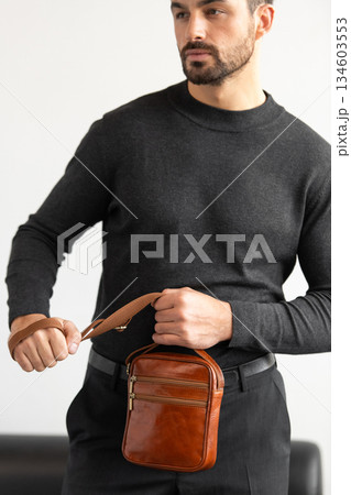 A stylish man in a dark grey crew neck sweater poses with a cognac leather crossbody bag against a minimalist white studio background. A stylish man in a dark grey crew neck sweater poses with a cognac leather crossbody bag against a minimalist white studio background. 134603553