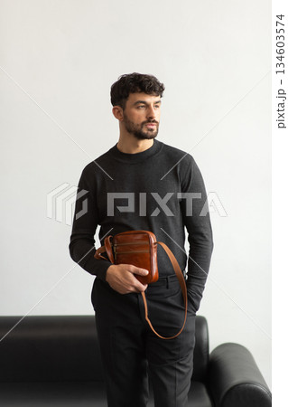 A stylish man in a dark grey crew neck sweater poses with a cognac leather crossbody bag against a minimalist white studio background. 134603574