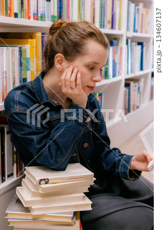 woman sits at shelves in library and reads book. education, scientific work. business person. 134607137
