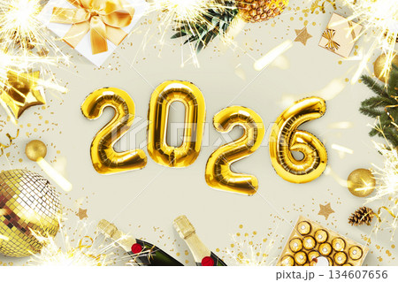 New Year 2026, creative idea. Golden balloons 2026 lie on a beige concrete table with fireplaces, candies, a golden disco ball, champagne and a Christmas tree, top view. Christmas card 2026 134607656