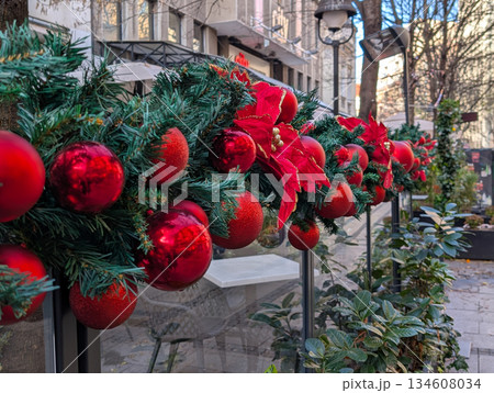 Close up of Christmas garland with red ornaments decorating an outdoor cafe fence in winter highlighting festive city details. Holiday atmosphere, urban decoration, seasonal design and cozy street 134608034