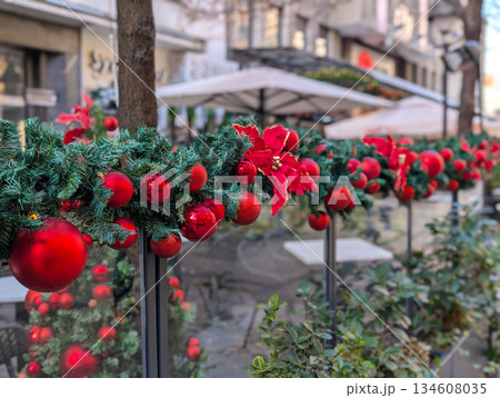 Festive Christmas decorations with red ornaments along a city cafe terrace on a winter street. Seasonal urban lifestyle, holiday mood, outdoor dining culture and decorative design in public space. Festive Christmas decorations with red ornaments along a city cafe terrace on a winter street. Seasonal urban lifestyle, holiday mood, outdoor dining culture and decorative design in public space. 134608035