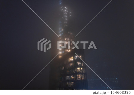 Futuristic Skyscraper Shrouded in Dense Night Fog. cinematic low-angle shot of a modern glass skyscraper piercing through thick night mist. Glowing office windows create a ethereal pattern, atmosphere Futuristic Skyscraper Shrouded in Dense Night Fog. cinematic low-angle shot of a modern glass skyscraper piercing through thick night mist. Glowing office windows create a ethereal pattern, atmosphere 134608350