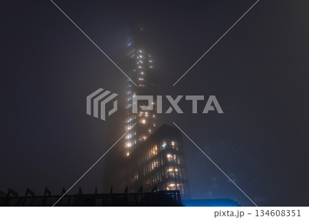 cinematic shot of a modern skyscraper disappearing into thick fog at night, featuring glowing office windows. Foggy night cityscape with illuminated office building windows glowing through dense mist. 134608351