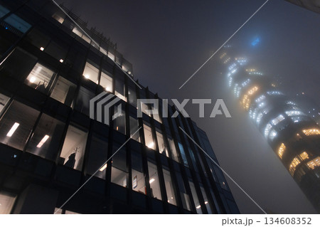 Moody cyberpunk-style architecture with a high-rise building glowing through dense urban mist and darkness. Foggy night cityscape with illuminated office building windows glowing Moody cyberpunk-style architecture with a high-rise building glowing through dense urban mist and darkness. Foggy night cityscape with illuminated office building windows glowing 134608352
