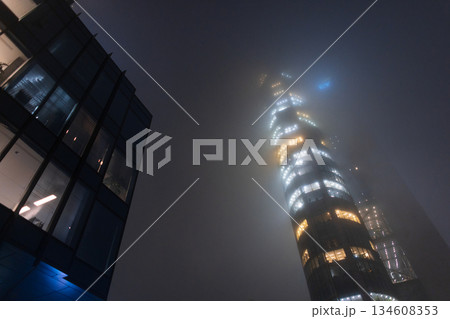Mysterious skyscraper shrouded in heavy night fog, creating an ethereal and atmospheric city scene. 134608353