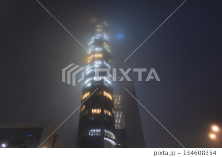 Low angle view of a contemporary glass office tower illuminated from within during a foggy night. Foggy night cityscape with illuminated office building windows glowing through dense mist. 134608354