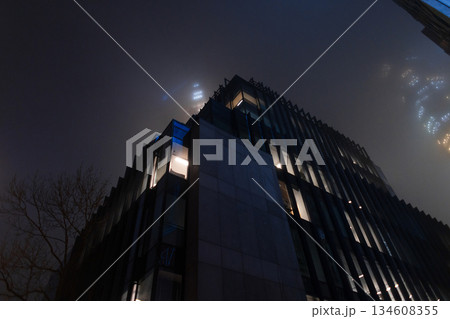 dark, dystopian urban landscape featuring a lonely skyscraper piercing through a thick layer of clouds. Foggy night cityscape with illuminated office building windows glowing through dense mist. dark, dystopian urban landscape featuring a lonely skyscraper piercing through a thick layer of clouds. Foggy night cityscape with illuminated office building windows glowing through dense mist. 134608355