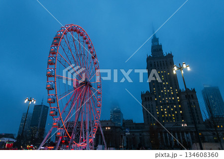 Dramatic low-angle view of an illuminated Ferris wheel in Warsaw at twilight. The glowing red attraction stands against a misty blue sky with the iconic Palace of Culture and Science in the background Dramatic low-angle view of an illuminated Ferris wheel in Warsaw at twilight. The glowing red attraction stands against a misty blue sky with the iconic Palace of Culture and Science in the background 134608360