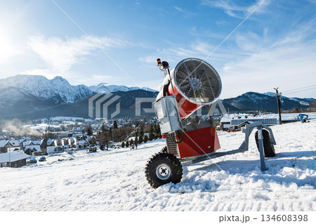 powerful, red mobile snow cannon sits on a bright, snow-covered ski slope with a stunning backdrop of the Tatra Mountains and a picturesque resort village. scene is captured on a clear, sunny winter 134608398