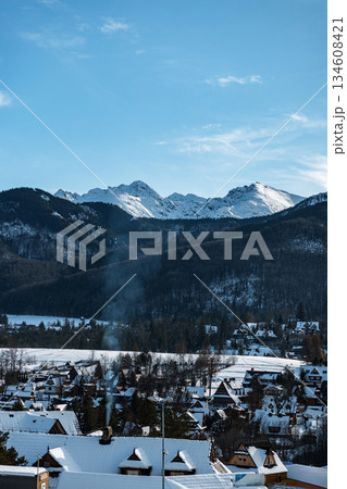 Breathtaking winter panorama of the picturesque alpine village nestled below the magnificent, sunlit, snow-capped Tatra Mountains against a clear blue sky. Winter holidays. Breathtaking winter panorama of the picturesque alpine village nestled below the magnificent, sunlit, snow-capped Tatra Mountains against a clear blue sky. Winter holidays. 134608421