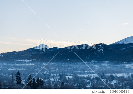 famous snow-capped peaks of the Tatra Mountains, including Giewont, stand above the winter resort town of Zakopane, Poland. Morning fog covers the valley, with sunlight breaking through a blue sky 134608435