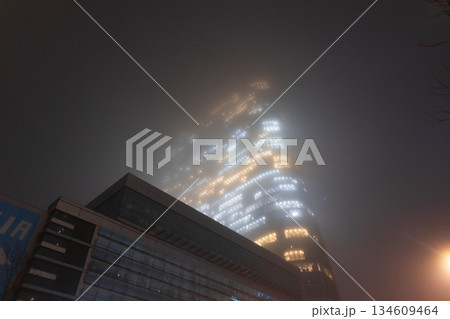 Warm electrical light from office cubicles contrasting with the cold, dark fog surrounding a glass skyscraper. Office lights blur into a luminous pattern, constant energy and power of a megacity Warm electrical light from office cubicles contrasting with the cold, dark fog surrounding a glass skyscraper. Office lights blur into a luminous pattern, constant energy and power of a megacity 134609464