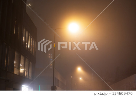 Foggy urban street at night illuminated by warm streetlights. Mist softens modern buildings and creates a moody, cinematic atmosphere, ideal for themes of mystery, solitude, weather, city life. 134609470