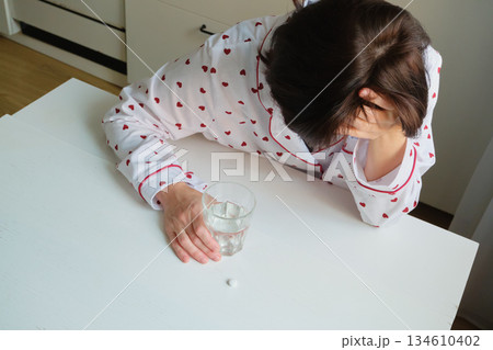 Woman wearing pajamas experiencing headache, sitting at table with medicine and water 134610402