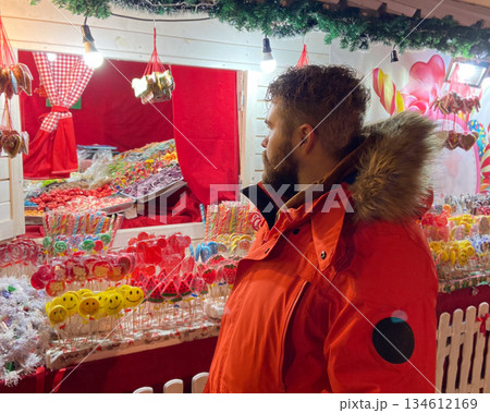 Man in winter parka choosing sweets at festive street stall. Holiday atmosphere, childhood nostalgia, colorful indulgence and joyful urban winter market experience. Man in winter parka choosing sweets at festive street stall. Holiday atmosphere, childhood nostalgia, colorful indulgence and joyful urban winter market experience. 134612169