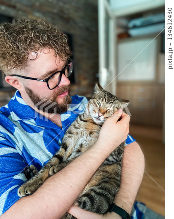 Man holding tabby cat in arms inside home interior. Emotional connection, pet companionship and calm domestic lifestyle expressing care, trust and everyday affection. 134612430