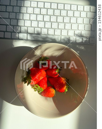 Fresh strawberries in bowl with sunlight on workspace table. Natural light mood, healthy snack choice and balanced workday lifestyle with minimal food composition. 134612438