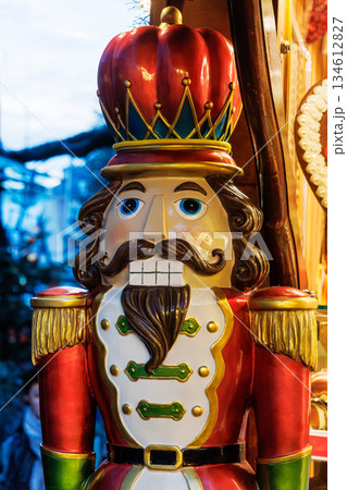 Christmas nutcracker figure at festive market stall. Wooden painted nutcracker in outdoor street setting with buildings and evening lights on blurred background Christmas nutcracker figure at festive market stall. Wooden painted nutcracker in outdoor street setting with buildings and evening lights on blurred background 134612827