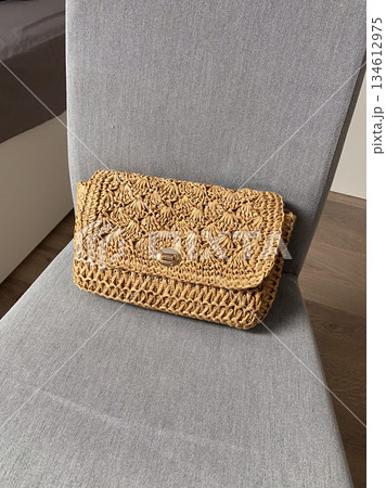 Straw clutch bag resting on a fabric chair indoors. Natural texture, handmade accessory, slow fashion, summer style and minimalist lifestyle 134612975