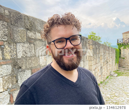 Smiling man portrait near historic stone wall outdoors. Positive travel emotion, relaxed vacation mood and authentic lifestyle experience within cultural heritage surroundings. Smiling man portrait near historic stone wall outdoors. Positive travel emotion, relaxed vacation mood and authentic lifestyle experience within cultural heritage surroundings. 134612978