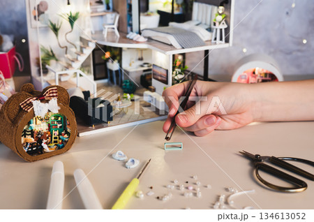 A girl uses tweezers to assemble decorations for a miniature dollhouse. A girl uses tweezers to assemble decorations for a miniature dollhouse. 134613052