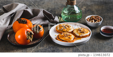 Delicious baked persimmon with cottage cheese, nuts and honey on a plate web banner Delicious baked persimmon with cottage cheese, nuts and honey on a plate web banner 134613072