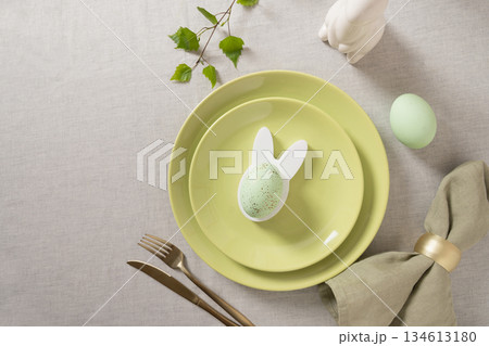 Stylish Easter table decor with bunny shaped egg holder, pastel egg, green plates and linen napkin on light textile background, flat lay composition. Stylish Easter table decor with bunny shaped egg holder, pastel egg, green plates and linen napkin on light textile background, flat lay composition. 134613180