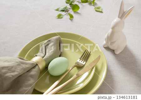 Minimal Easter place setting with pastel egg, green ceramic plates, golden cutlery and white bunny figurine on neutral tablecloth, spring holiday concept. Minimal Easter place setting with pastel egg, green ceramic plates, golden cutlery and white bunny figurine on neutral tablecloth, spring holiday concept. 134613181