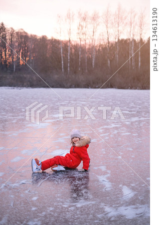 A child laughing from his fall lies on the ice surface of a frozen lake. Ice skating on a frosty day in nature 134613190