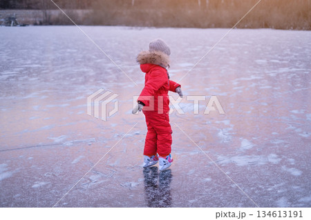 A girl in a bright red winter jumpsuit skates on the ice rink of a frozen lake at sunset 134613191