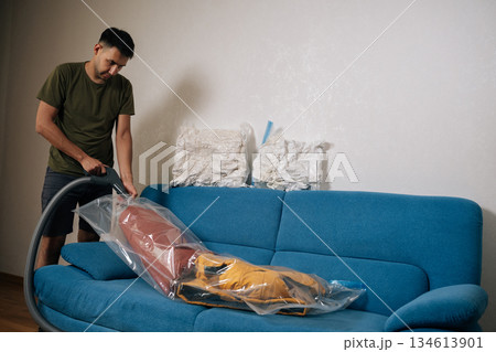 Focused man using vacuum cleaner to remove air from vacuum storage bag with clothes and placing it on sofa in living room, organizing seasonal wardrobe for winter. Concept of household chores. Focused man using vacuum cleaner to remove air from vacuum storage bag with clothes and placing it on sofa in living room, organizing seasonal wardrobe for winter. Concept of household chores. 134613901