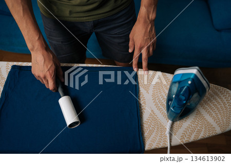 Top view of man using sticky lint roller to remove dust and lint from blue t-shirt on ironing board, with steam iron nearby, preparing to iron and care for clothes at home. Concept of housekeeping Top view of man using sticky lint roller to remove dust and lint from blue t-shirt on ironing board, with steam iron nearby, preparing to iron and care for clothes at home. Concept of housekeeping 134613902