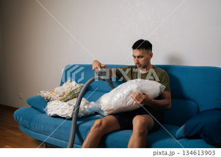 Focused young man sitting on sofa using vacuum cleaner to suck air out of storage bag containing bedding, reducing storage space and protecting from dust and moisture. Concept of household chores. Focused young man sitting on sofa using vacuum cleaner to suck air out of storage bag containing bedding, reducing storage space and protecting from dust and moisture. Concept of household chores. 134613903