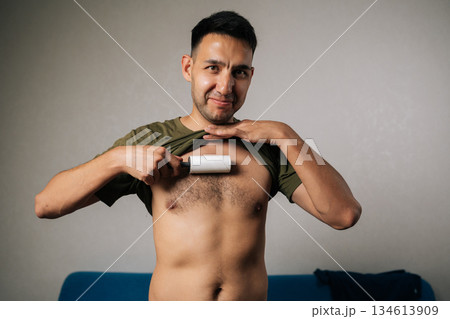 Joyful young man lifting shirt and using sticky lint roller to remove hair and fluff from chest at home, showcasing humorous approach to personal grooming, smiling looking at camera. Joyful young man lifting shirt and using sticky lint roller to remove hair and fluff from chest at home, showcasing humorous approach to personal grooming, smiling looking at camera. 134613909