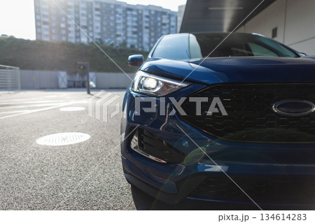Modern automobile in metallic blue finish parked in city, close-up of front bumper and lights. Modern transportation concept, blue SUV parked on asphalt street with sunlight reflections. 134614283
