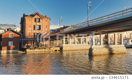 Porvoo, Finland - December 08, 2020: Historic red brick building stands by the river, with a modern bridge overhead, showcasing architectural contrast and scenic waterfront views Porvoo, Finland - December 08, 2020: Historic red brick building stands by the river, with a modern bridge overhead, showcasing architectural contrast and scenic waterfront views 134615171