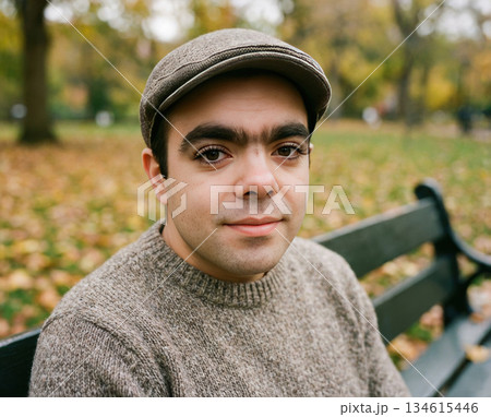 young adult sits on a park bench wearing a sweater and cap. Leaves cover the ground around them. The sun shines bright in a park setting with trees in the background. 134615446