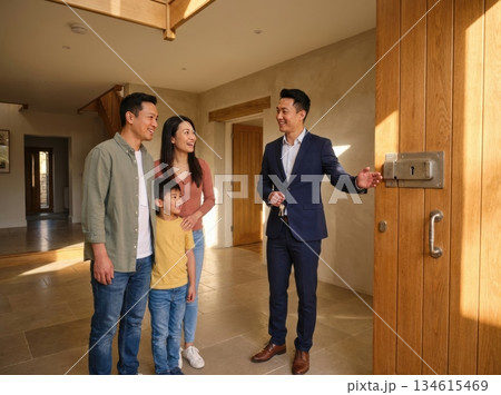 realtor stands at the entrance of a house showing it to an Asian couple and their child. They are smiling and interacting in a bright and spacious living area. 134615469