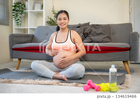 Pregnant asian woman massaging her bare belly while sitting on rubber mat in the house's living room 134619466