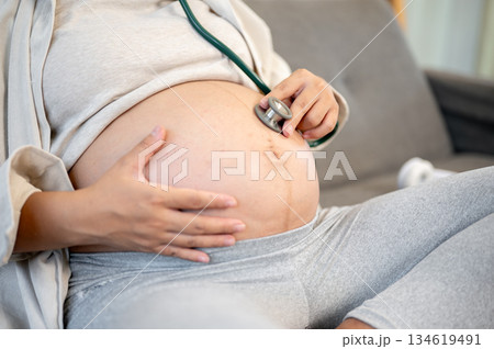 Close up of pregnant woman using stethoscope on belly listening to fetal heartbeats sitting on sofa 134619491