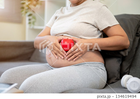 Pregnant asian woman making a heart with fingers over red ball on bare belly while sitting on sofa. Pregnant asian woman making a heart with fingers over red ball on bare belly while sitting on sofa. 134619494
