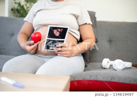 Pregnant asian woman holding a rubber heart and ultrasound pictures on her belly sitting on the sofa 134619497