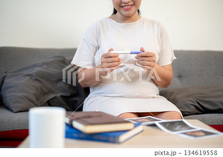 Pregnant asian woman is smiling and looking at pregnancy test kit in hand sitting on sofa in house. Pregnant asian woman is smiling and looking at pregnancy test kit in hand sitting on sofa in house. 134619558