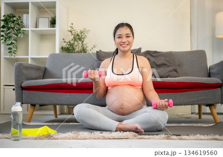 Pregnant asian woman with bare belly smiling as lifting dumbbell while sitting on mat in living room Pregnant asian woman with bare belly smiling as lifting dumbbell while sitting on mat in living room 134619560