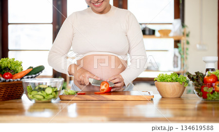 Close up of pregnant woman holding knife cutting tomato on chopping board making salad in kitchen 134619588