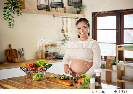 Pregnant asian woman smiling as she cutting cucumbers with knife on chopping board in the kitchen. Pregnant asian woman smiling as she cutting cucumbers with knife on chopping board in the kitchen. 134619589