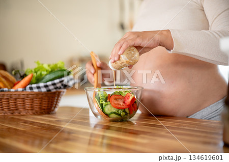 Close up of pregnant woman putting mayonnaise into glass bowl making a vegetables salad in kitchen. Close up of pregnant woman putting mayonnaise into glass bowl making a vegetables salad in kitchen. 134619601