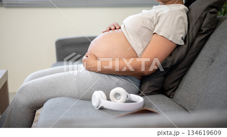Close up of pregnant asian woman holding bare belly in both hands sitting leaning on sofa in house. 134619609