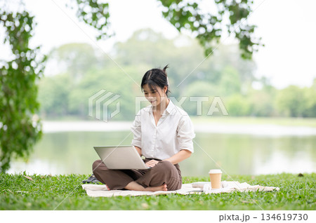 Asian woman in white shirt looking or using her laptop while sitting on picnic mat laying on grass. 134619730